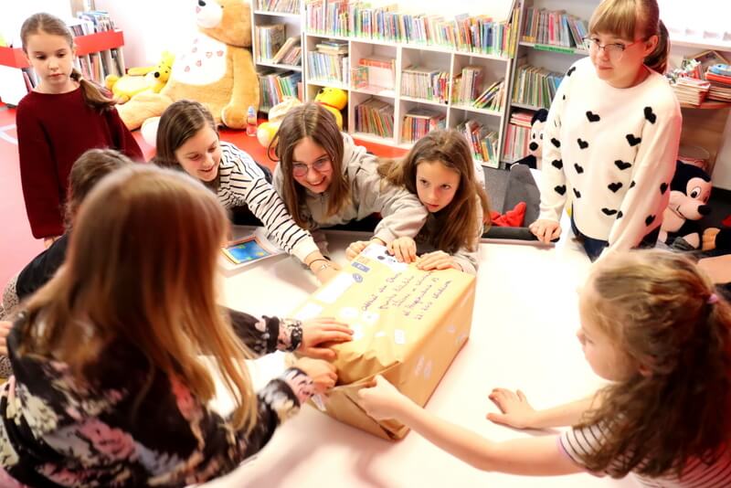 Grupa dzieci siedzi wokół stołu na którym stoi ogromna paczka, próbując ją rozpakować. Tło stanowią regały z książkami.