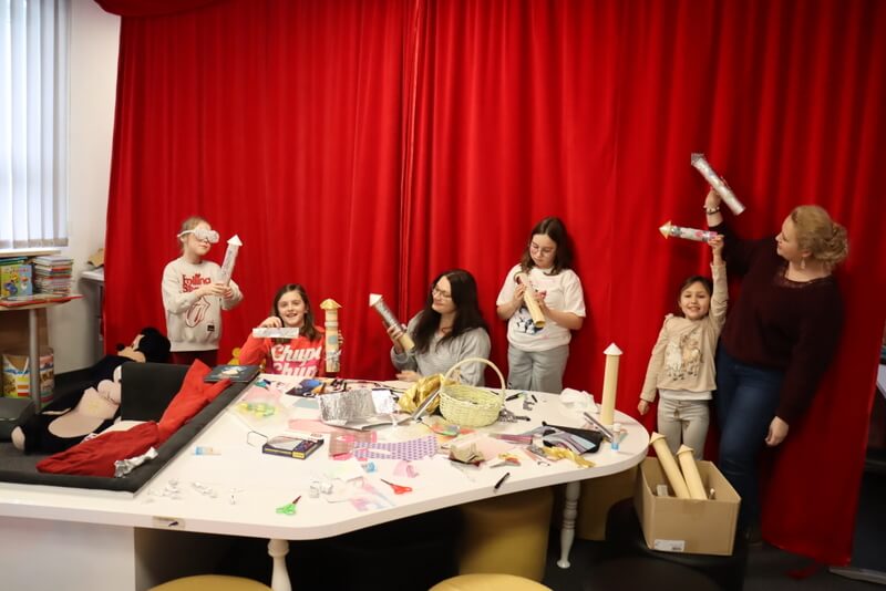 Dzieci wraz z prowadzącymi siedzą wokół stołu z książkami. Z magicznego kuferka losują ciekawostki związane z tematem dzisiejszych zajęć. W tle widnieją wyeksponowane portrety słynnych polaków.