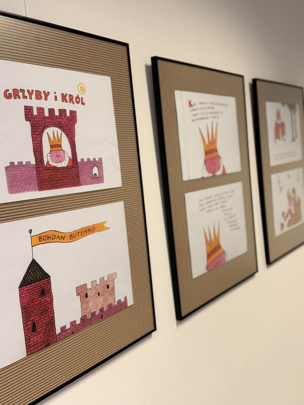 Trzy oprawione w czarne ramy plansze wiszące na białej ścianie w galerii, prezentujące ilustracje książkowe Bohdana Butenki z serii „Grzyby i król”. Na pierwszym planie widać rysunki zamku, króla w koronie oraz flagę z nazwiskiem autora. Pozostałe dwie ramki usytuowane są w głąb kadru w perspektywie.