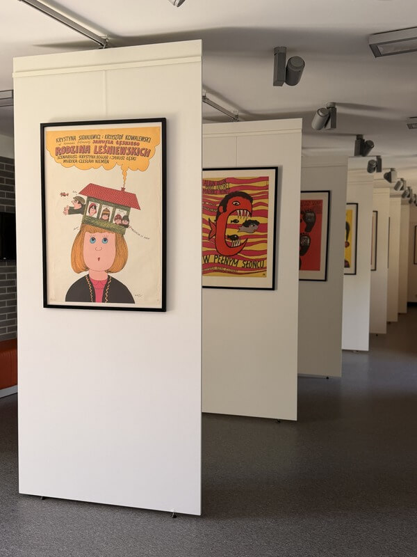 Wnętrze galerii ze stojącymi, białymi panelami wystawowymi, na których wiszą oprawione plakaty. Na pierwszym planie wyraźnie widać plakat do filmu „Rodzina Leśniewskich”. Dalsze panele tworzą perspektywiczny rząd w głąb sali, prezentując kolejne kolorowe grafiki.