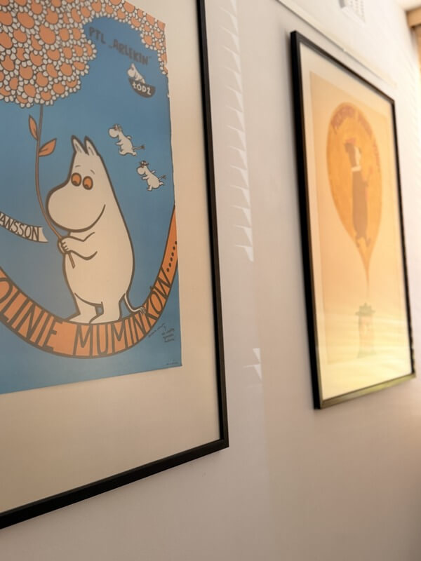 Zbliżenie na oprawiony plakat teatralny do spektaklu „W Dolinie Muminków” w Państwowym Teatrze Lalek „Arlekin” w Łodzi. Plakat przedstawia postać Muminka na niebieskim tle, trzymającego dużą gałąź z pomarańczowym kwiatem. W tle, po prawej stronie ściany, widać fragment kolejnego oprawionego plakatu w ciepłych tonacjach.