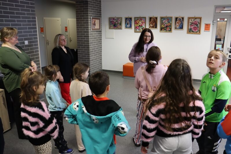 Galeria Na Piętrze. Na holu stoi grupka dzieci w różnym wieku, dwie panie – opiekunki i słuchają prowadzącej (J. Orzeszko). Na ścianach wiszą kolorowe obrazy wykonane haftem diamentowym.